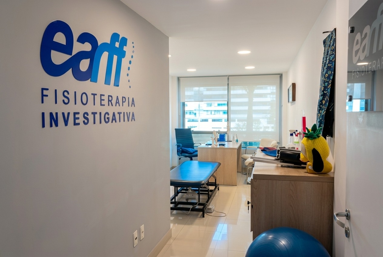 Corredor de entrada da unidade EARFF Fisioterapia Investigativa Freguesia, parede cinza clara com logo azul da marca em letras grandes, piso porcelanato claro, sala com maca de fisioterapia azul, mesa de madeira e boneco de pelúcia amarelo, janela com persianas ao fundo, iluminação natural suave, ambiente clean e moderno, fotografia arquitetônica realista