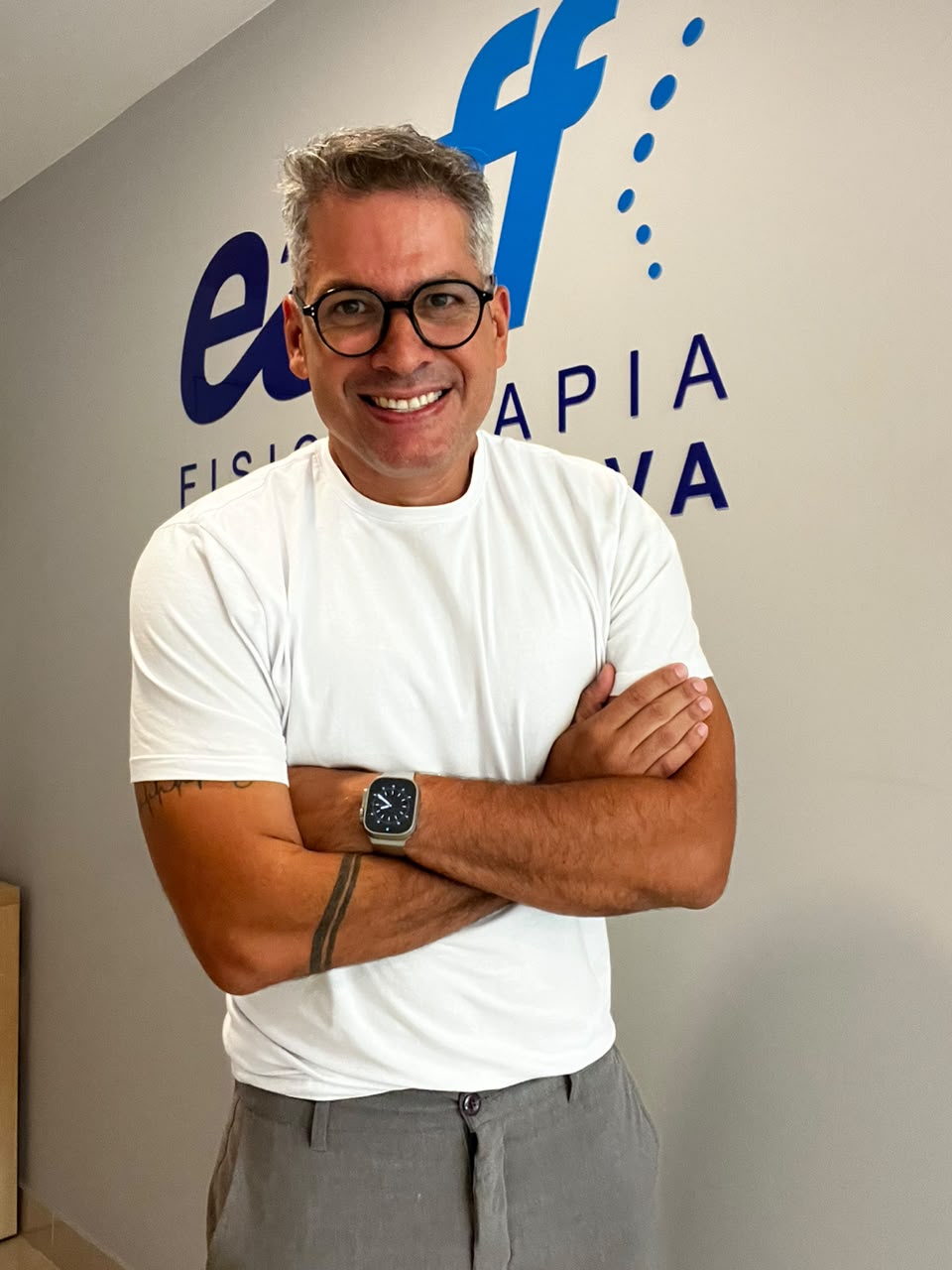 Retrato profissional do Dr. Fernando Capristano, fisioterapeuta da EARFF, camiseta branca, óculos de grau, braços cruzados, sorriso confiante, parede com logo EARFF Fisioterapia Investigativa ao fundo, fotografia corporativa realista