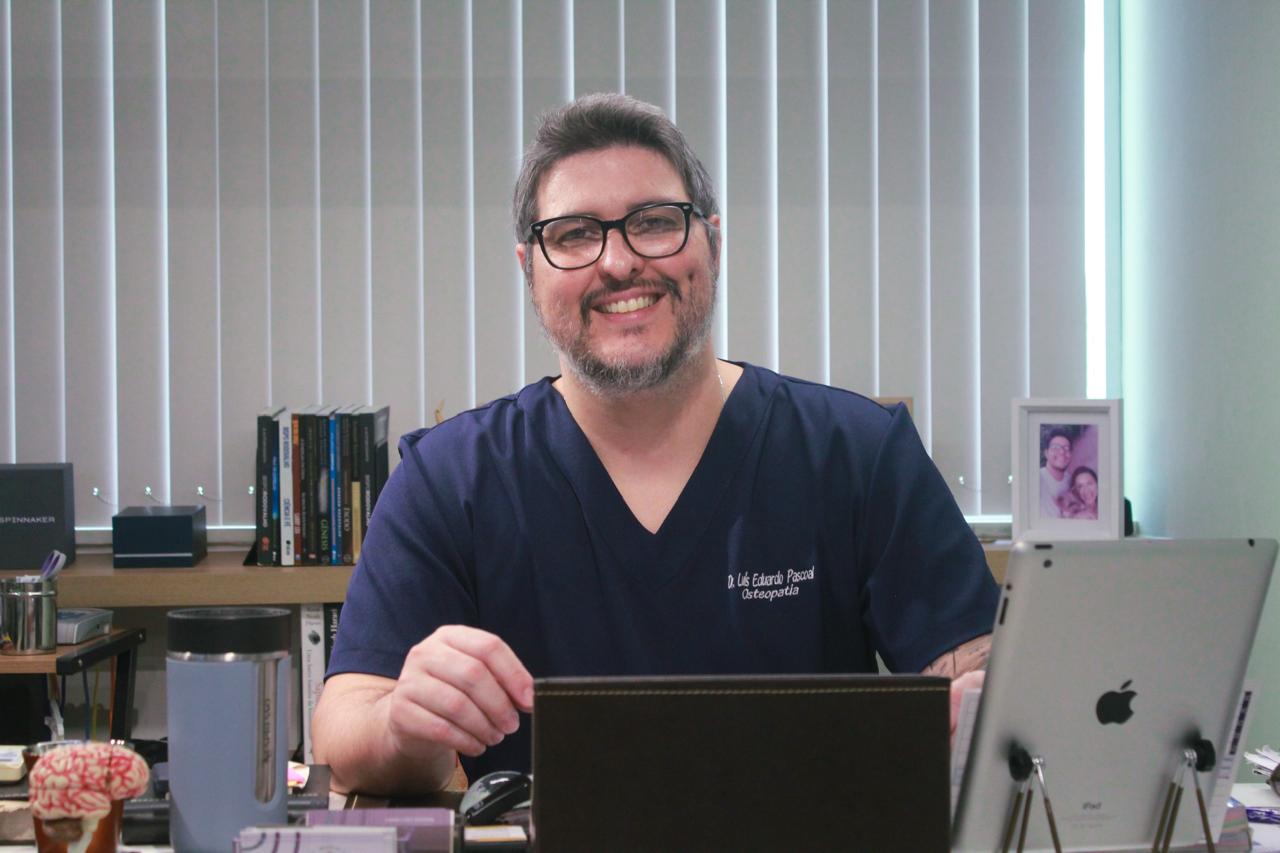 Retrato profissional do Dr. Luís Eduardo Pascoal, fisioterapeuta da EARFF, jaleco azul-marinho com bordado do nome, óculos de grau, sorriso confiante, sentado em consultório clínico com persianas ao fundo, fotografia corporativa realista