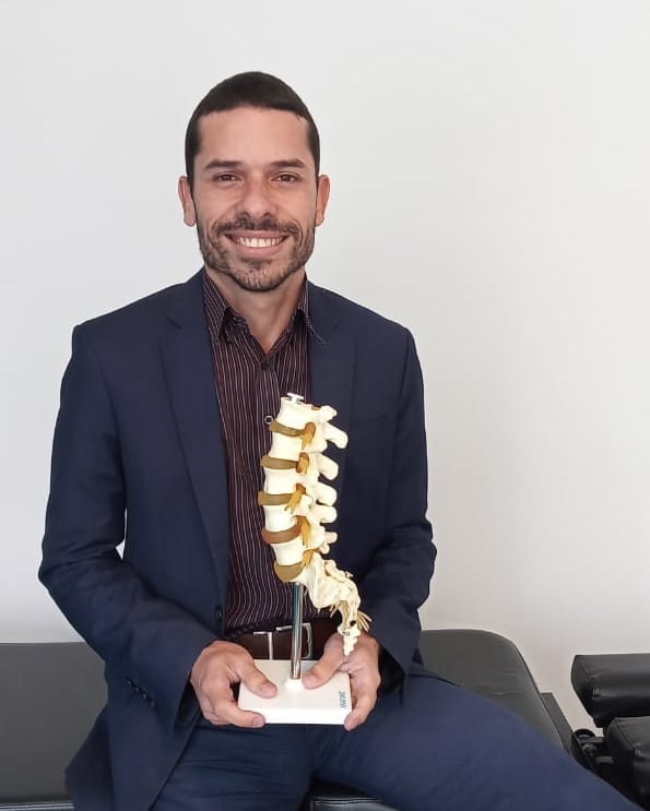 Retrato profissional do Dr. Angel Mello, fisioterapeuta da EARFF, terno azul-marinho, camisa listrada, segurando modelo anatômico de coluna vertebral, sorriso confiante, fundo neutro claro, fotografia corporativa realista